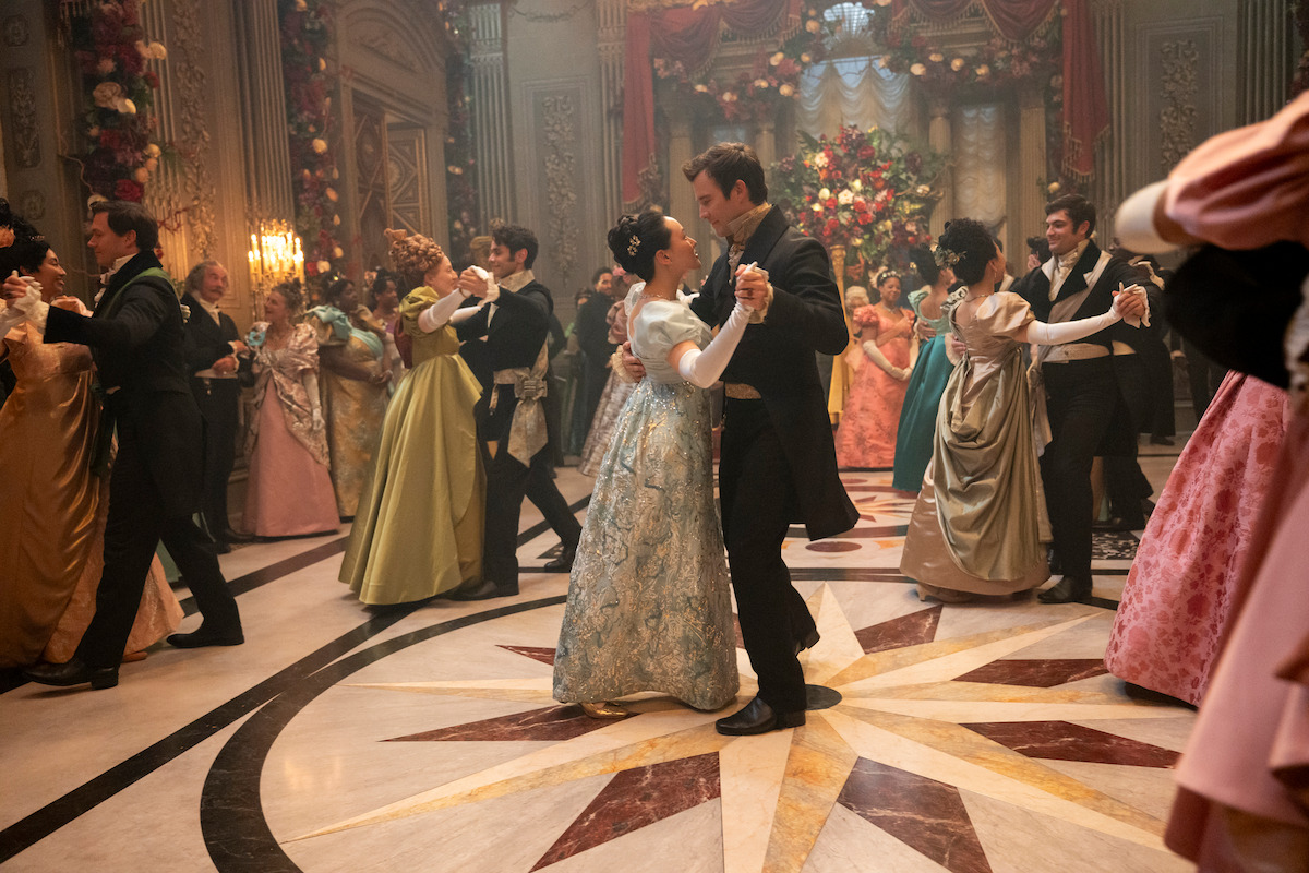 Elegant ballroom scene with couples in period costumes dancing on a polished marble floor, surrounded by ornate decorations, grand columns, and floral arrangements in a lavish, historical setting.