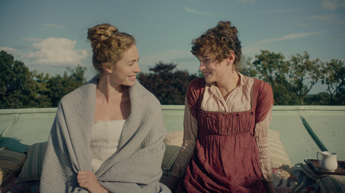 Two women in historical clothing sit outside on a rooftop, smiling at each other with greenery and blue sky in the background. Pillows and a cup are nearby, creating a relaxed, intimate setting.