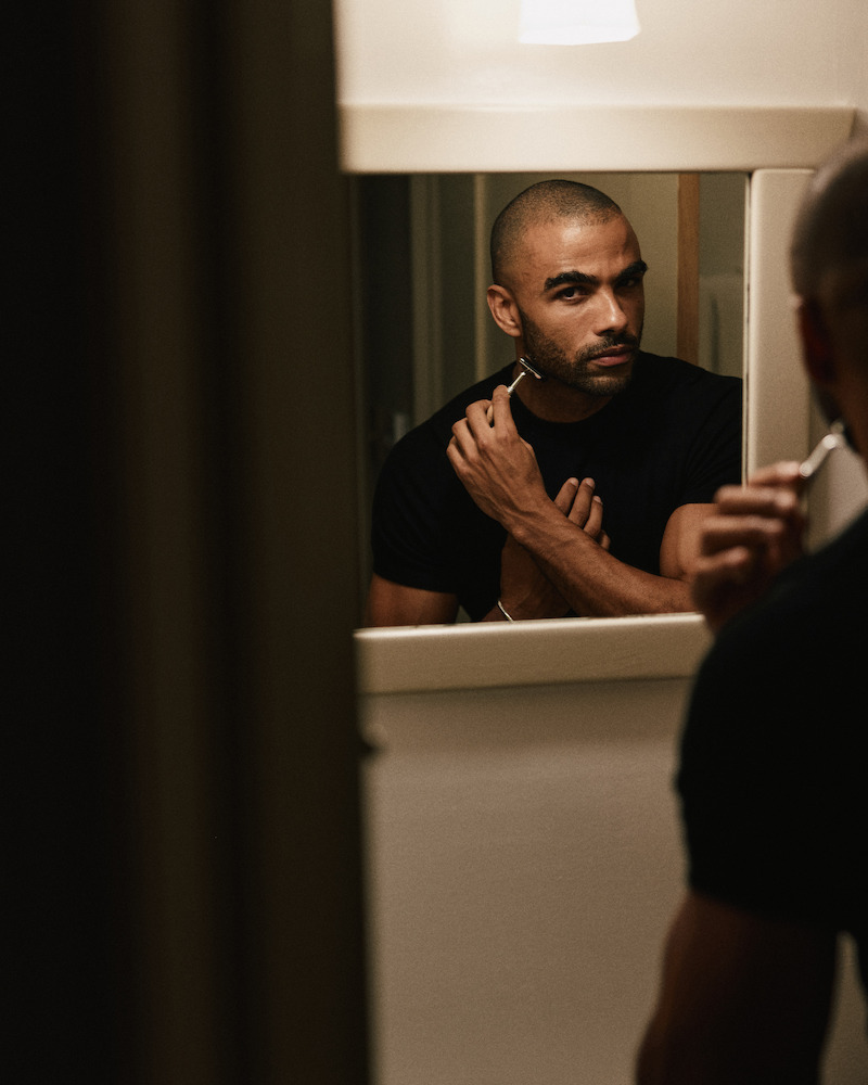 Man with a shaved head in a dark T-shirt stands in front of a bathroom mirror under warm lighting, shaving his face with a razor, focused on his reflection in a home setting.