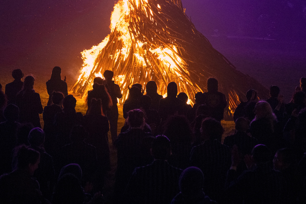 Silhouettes of a crowd gathered around a large bonfire at night in ‘Wednesday’ Season 2.