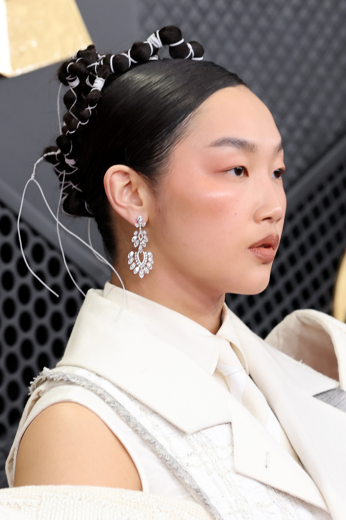 Woman with intricate updo and large jeweled earrings, dressed in elegant white attire, poses against a modern, geometric background with textured, dark panels.