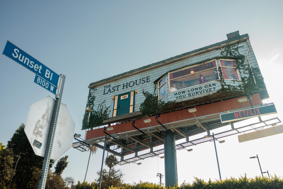 Billboard designed as a house with “The Last House” and “How long can you survive?” promoting Netflix, surrounded by greenery, located at the intersection of Sunset Blvd and 8100 W, under a clear sky.