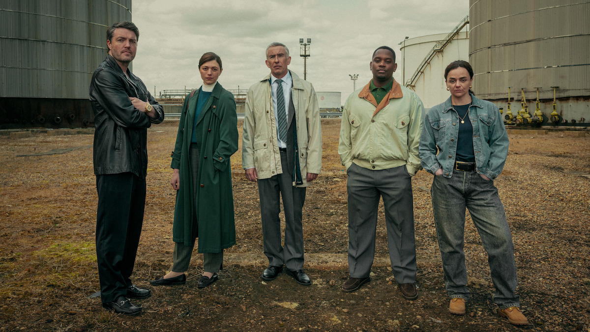 Five people stand in a gritty industrial outdoor setting between large storage tanks, dressed in various jackets and coats, facing the camera on a cloudy day, suggesting a serious or dramatic atmosphere.