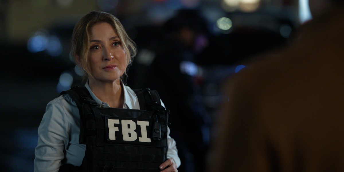 Woman wearing an FBI vest stands outdoors at night, facing a person out of focus in the foreground, with blurred emergency vehicles and personnel in the background, suggesting an active investigation scene.