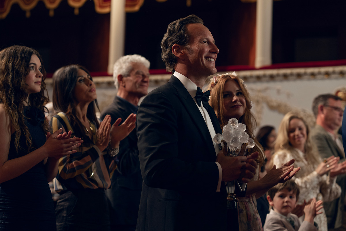 A man in a tuxedo and a woman with a flower crown sit in a dimly lit theater, surrounded by an attentive audience, creating a formal, relaxed, and engaged atmosphere.