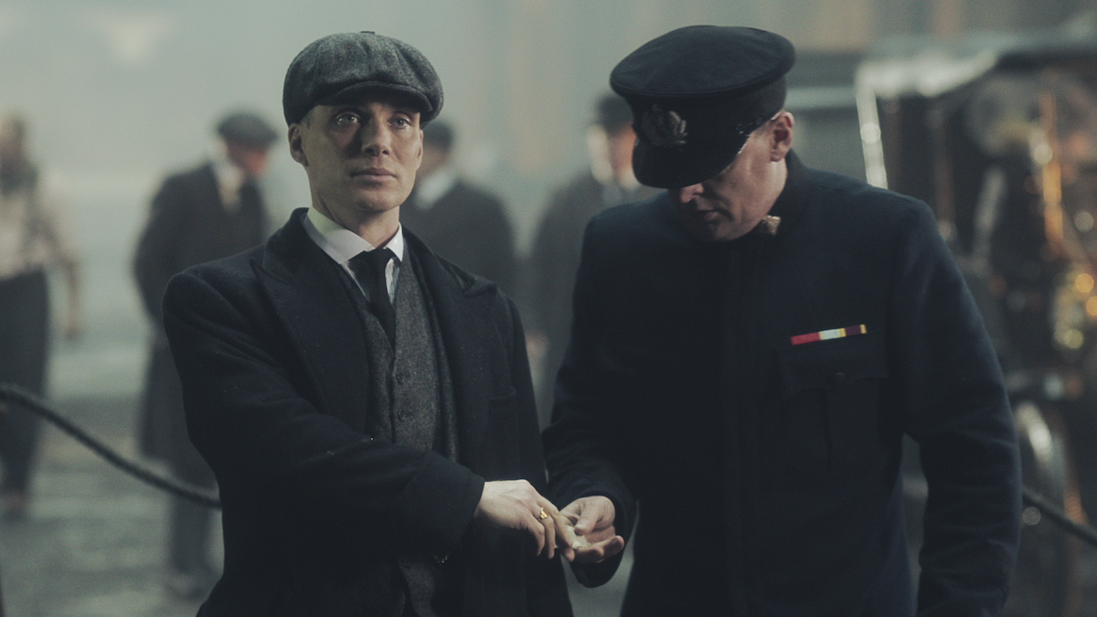 A man in a suit and hat hands a man in a uniform money as he looks off into the distance.