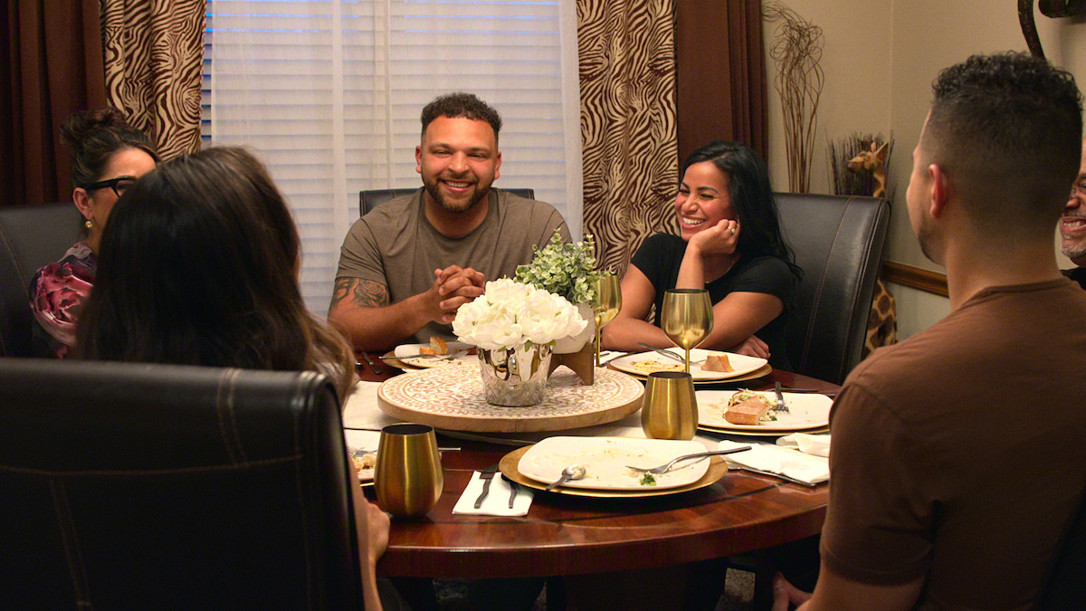 Group of people sitting around a dining table, smiling and talking, with plates and gold cups on the table in a warmly lit, cozy dining room setting.