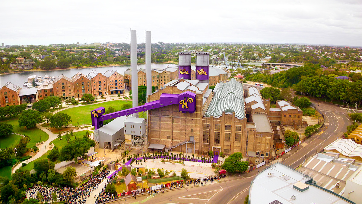 Large brick factory-like building with Wonka-themed decorations, purple banners, and smokestacks. Surrounded by green park, river, and other brick buildings. People queue outside on a cloudy day in a city environment.