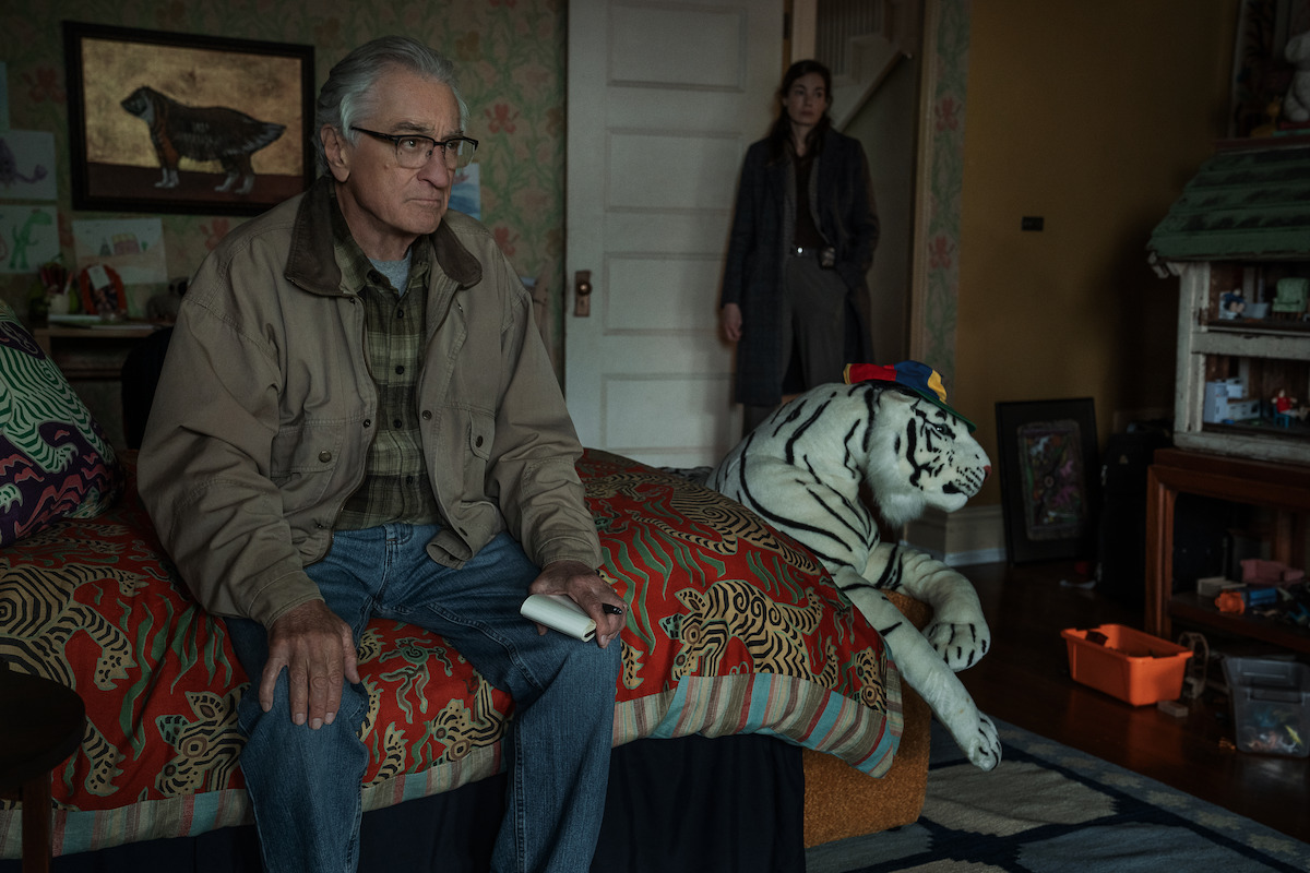 Older man sits on patterned bed holding remote in cozy, cluttered room with wallpaper, stuffed white tiger toy, and woman standing by the door in background. Warm lighting and eclectic decor give a homey feel.
