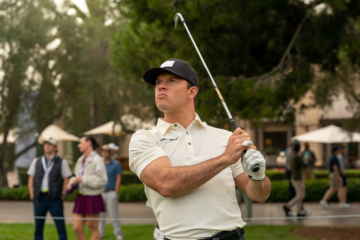 Man in white golf shirt and black cap holding a golf club after a swing on a golf course, with spectators and trees in the background on a cloudy day.