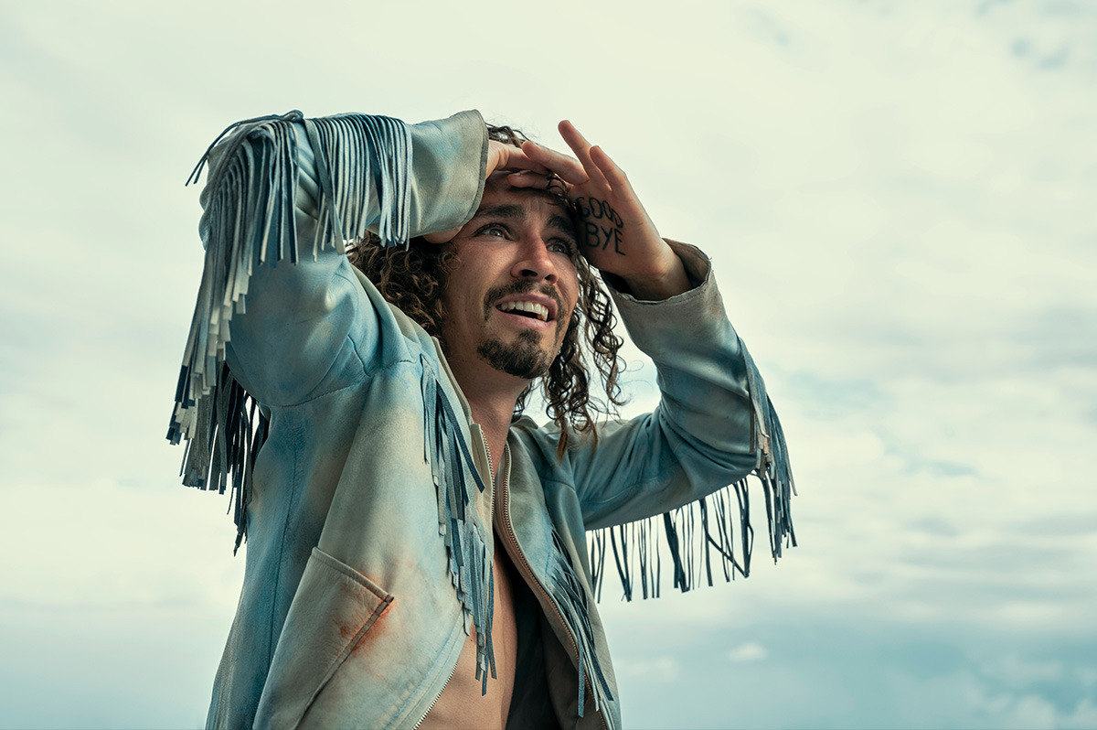  A man with curly hair wearing a fringed denim jacket, looking out into the distance.