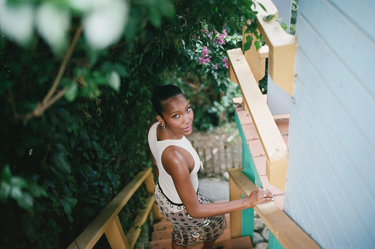Woman in a sleeveless top and patterned skirt stands on outdoor wooden stairs, surrounded by lush green foliage and flowers, looking up towards the camera next to a light blue building.