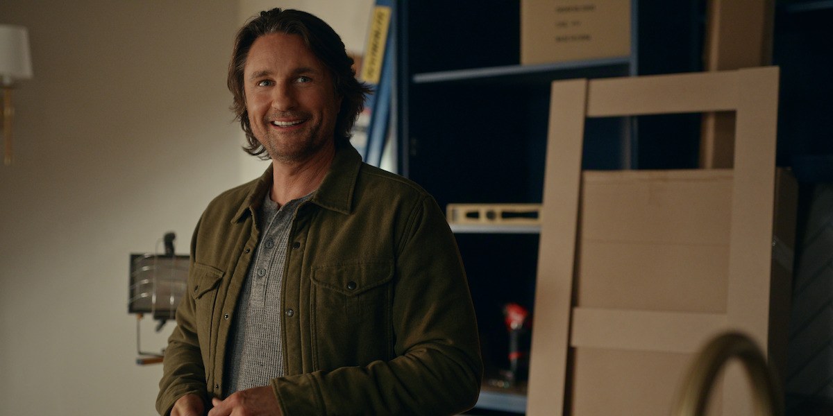 A man with long hair and a beard smiles while standing indoors in a casual setting, wearing a green jacket and grey shirt, with shelving and boxes in the background.