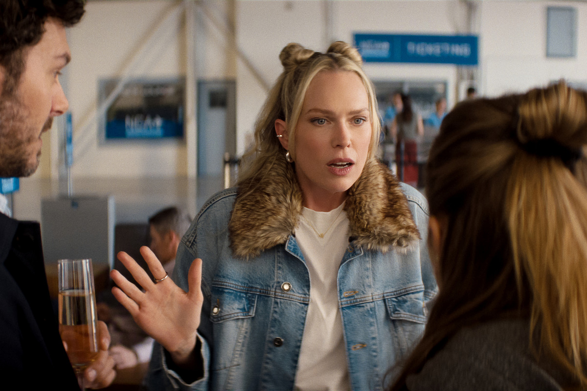 A woman in a denim jacket with a fur collar gestures while talking to two people at an indoor airport terminal, with signs and passengers visible in the background.