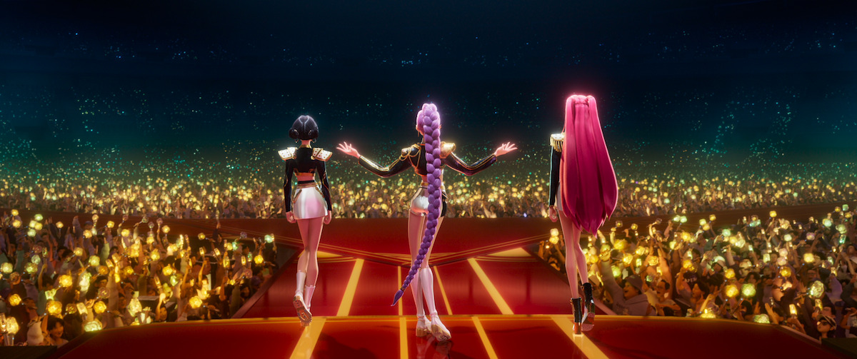 Three animated female performers with colorful hair stand on a large stage facing a cheering crowd holding glowing lights in an arena, suggesting a vibrant concert or live performance scene.