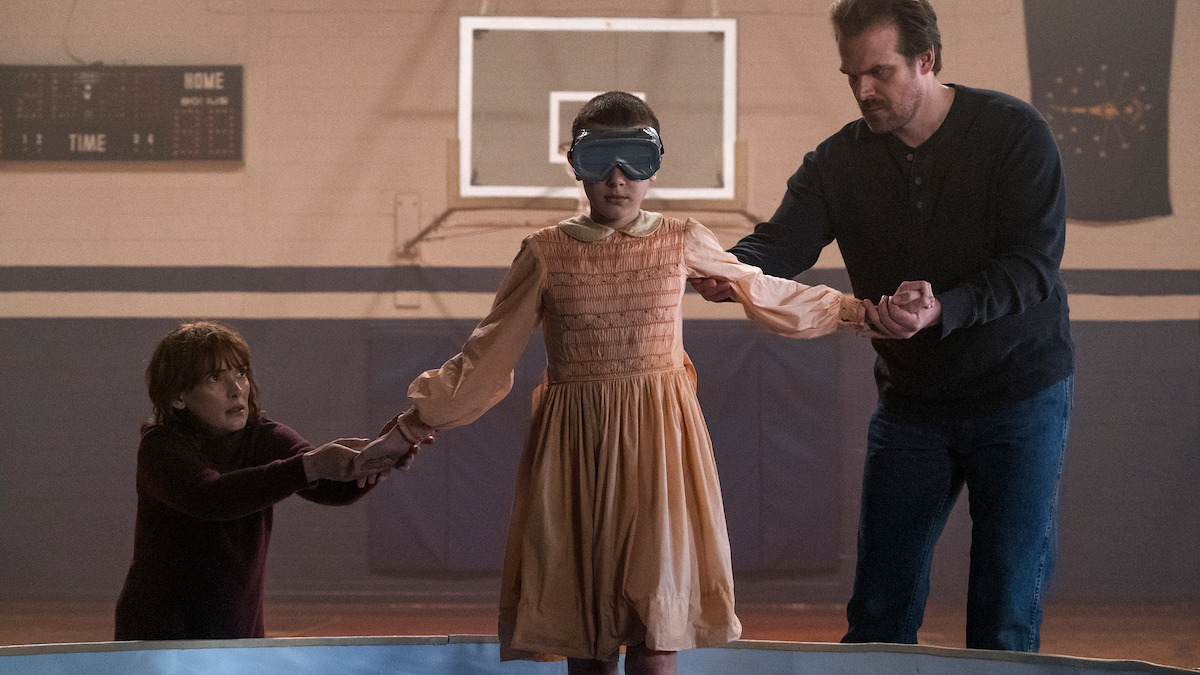 A girl in a dress and blindfold stands in a gym, holding hands with a man and a boy. The mood is tense and focused, with a basketball hoop, scoreboard, and tank visible in the background.