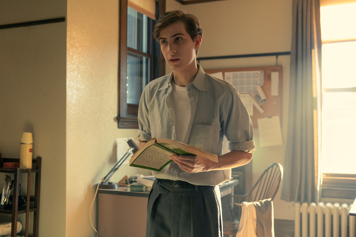 Young person in casual clothes standing in a warmly lit room, holding an open book. The environment appears to be a study or office, with a desk, chair, and bulletin board visible, and sunlight streaming through the window.