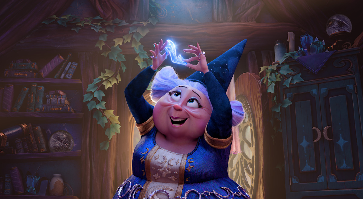 A cheerful animated female wizard in a blue robe and hat holds a glowing magical object above her head inside a cozy, book-filled, ivy-adorned room with wooden furniture and mystical decorations.