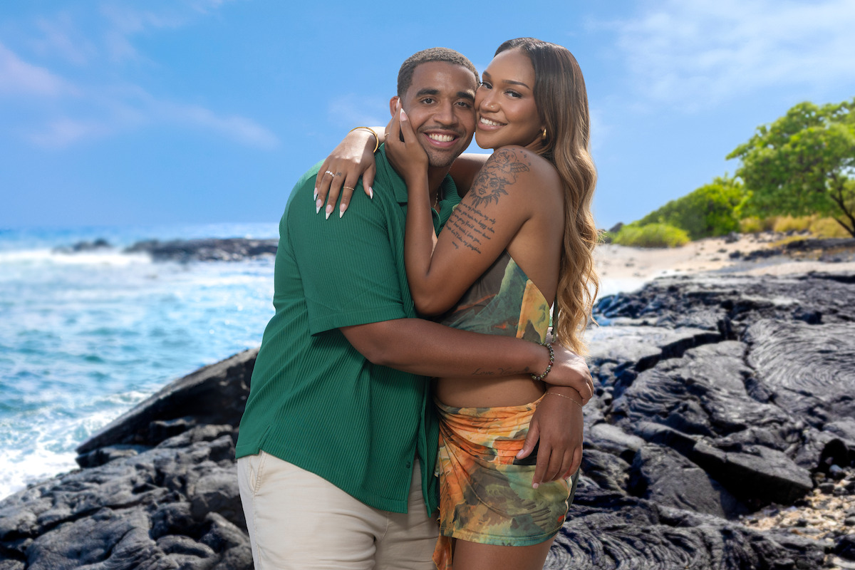 Smiling couple embracing on a rocky beach with ocean waves, blue sky, and lush green trees in the background, enjoying a sunny day in a tropical outdoor setting.
