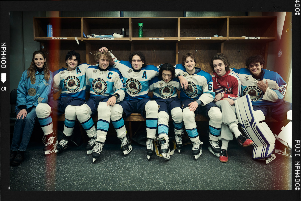 A group of young hockey players in blue and white uniforms sits on a bench, smiling and posing for a team photo in a locker room with wooden cubbies behind them.