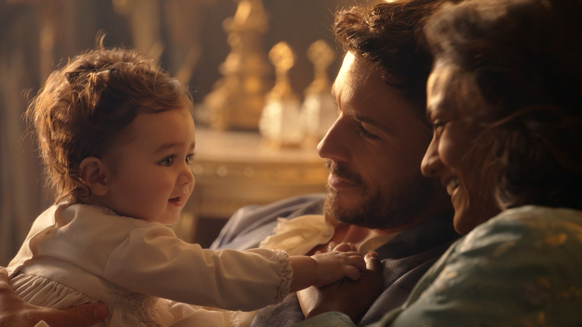 A smiling baby in light clothing is held by two adults, likely parents, sharing a joyful, intimate moment together in a warmly lit, elegant room with decorative items in the background.