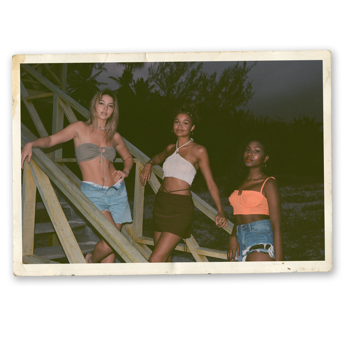 Three women posing on wooden stairs outdoors at dusk, dressed in summer tops and shorts, with trees and dark sky in the background, vintage photo style.