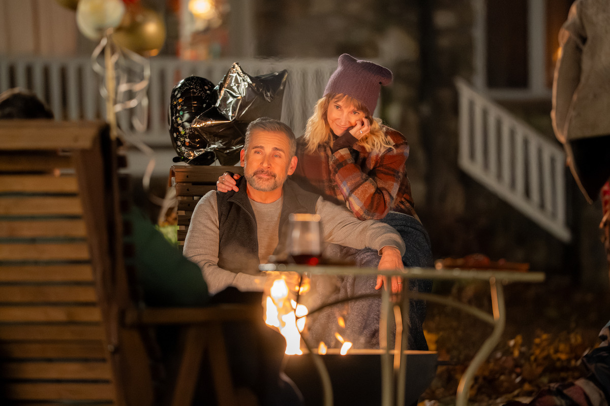 Man and woman sitting by an outdoor firepit at night, with cozy autumn clothing, balloons, and warm lighting in a backyard gathering setting.