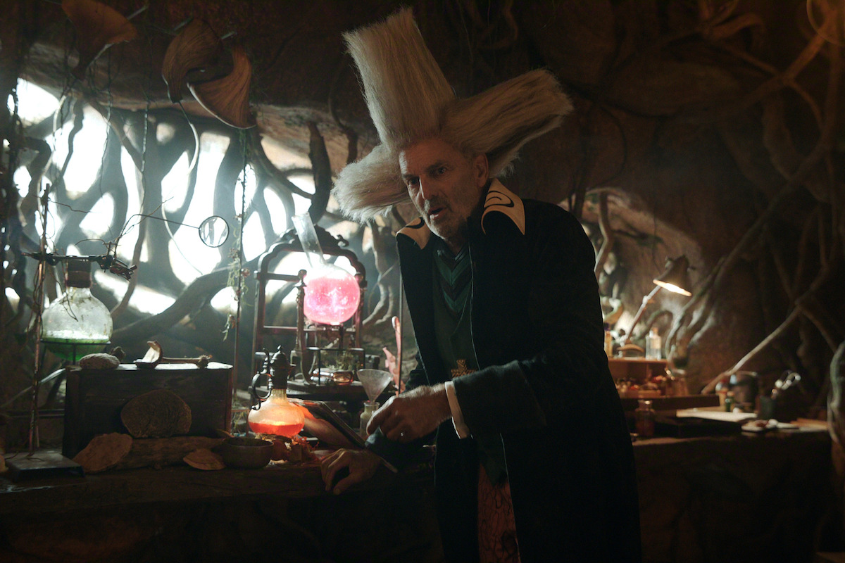 Man with tall, wild hair in a fantastical laboratory filled with glowing potions, twisted roots, and strange scientific equipment, appearing to conduct an experiment in a mystical, dimly lit environment.