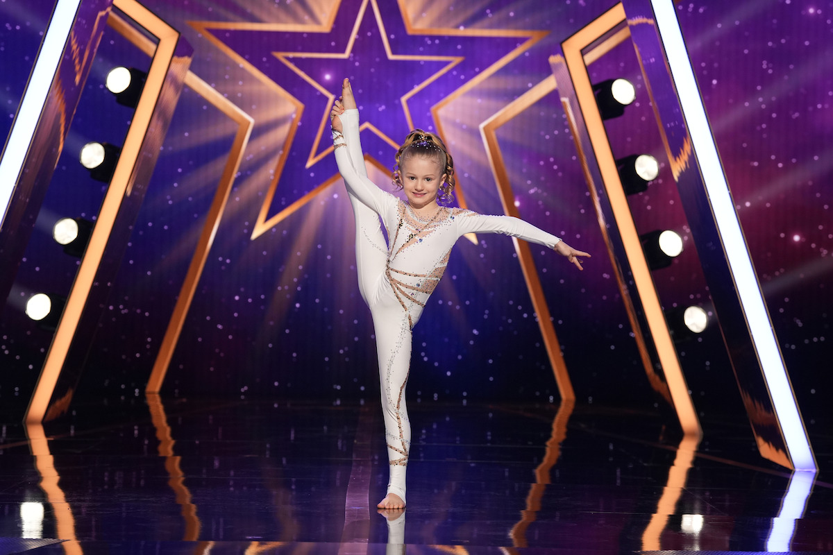 Kaylen Fairchild in a white leotard performing a high leg stretch on a shiny stage with purple starry background and theatrical lighting, likely at a talent or dance competition.