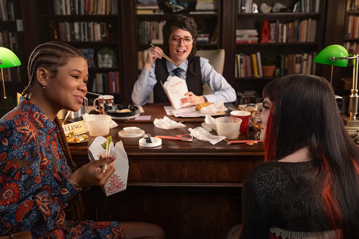 Three people sharing takeout food and laughing in a cozy office with a large wooden desk, green lamps, paperwork, and a bookshelf in the background, creating a relaxed and friendly atmosphere.