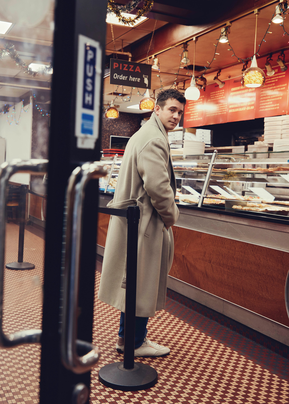 Man in a long beige coat standing in line at a pizzeria counter with pizzas and pizza boxes, decorated with lights and a patterned floor, looking toward the camera.
