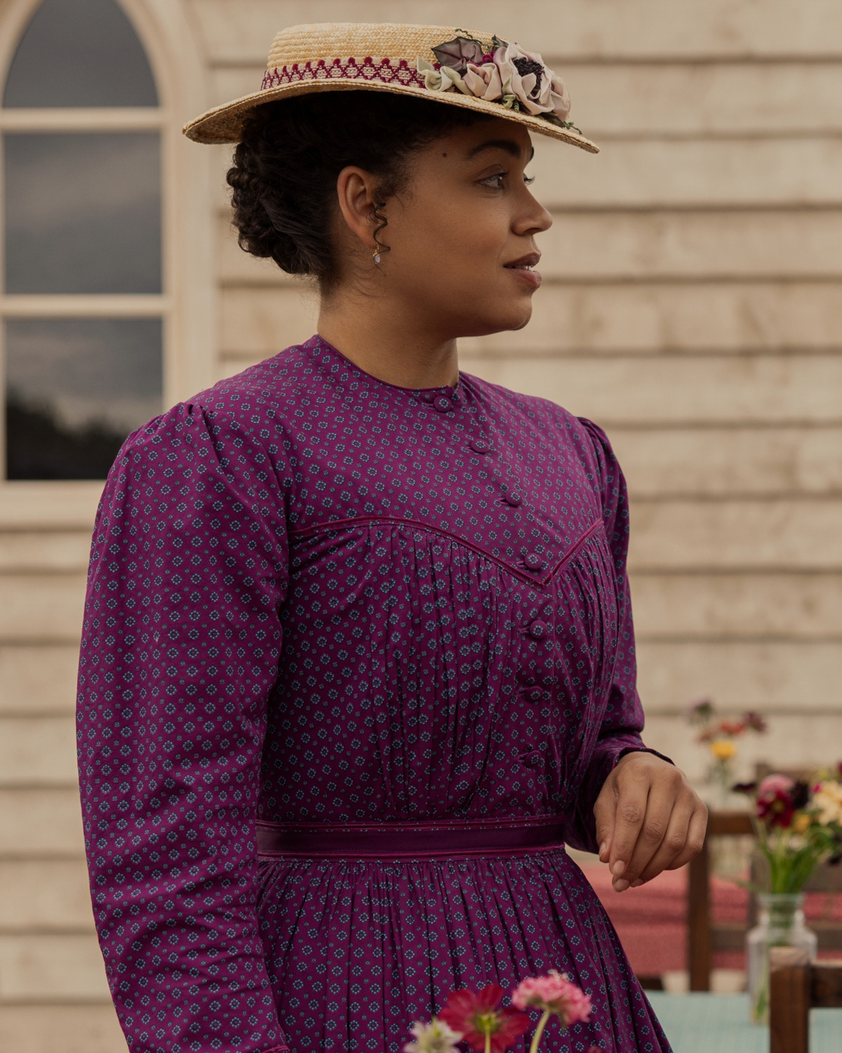 Woman in a purple patterned dress and straw hat with flowers, standing outdoors by a wooden building, with a window and flowers in the background, looking to the side in daylight.