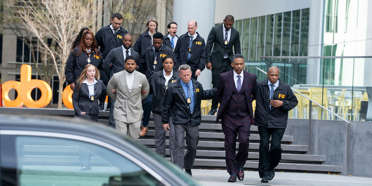 A group of FBI agents escorting two men in suits down the steps outside a modern building in an urban setting, with a car partially visible in the foreground and office seating in the background.