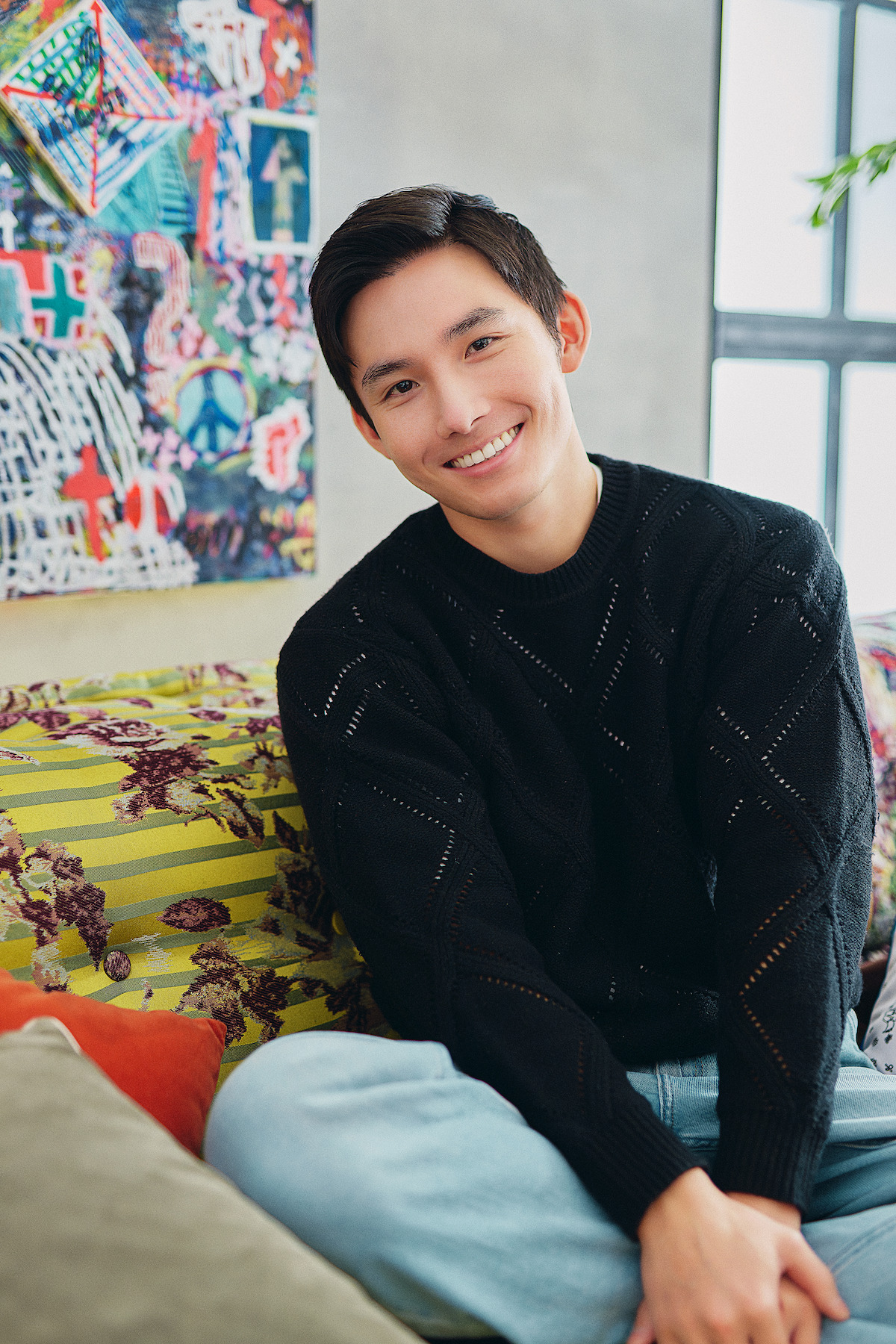 Young man smiling and sitting on a colorful couch with floral patterns in a modern, bright living room with abstract artwork on the wall, creating a relaxed and cheerful atmosphere.