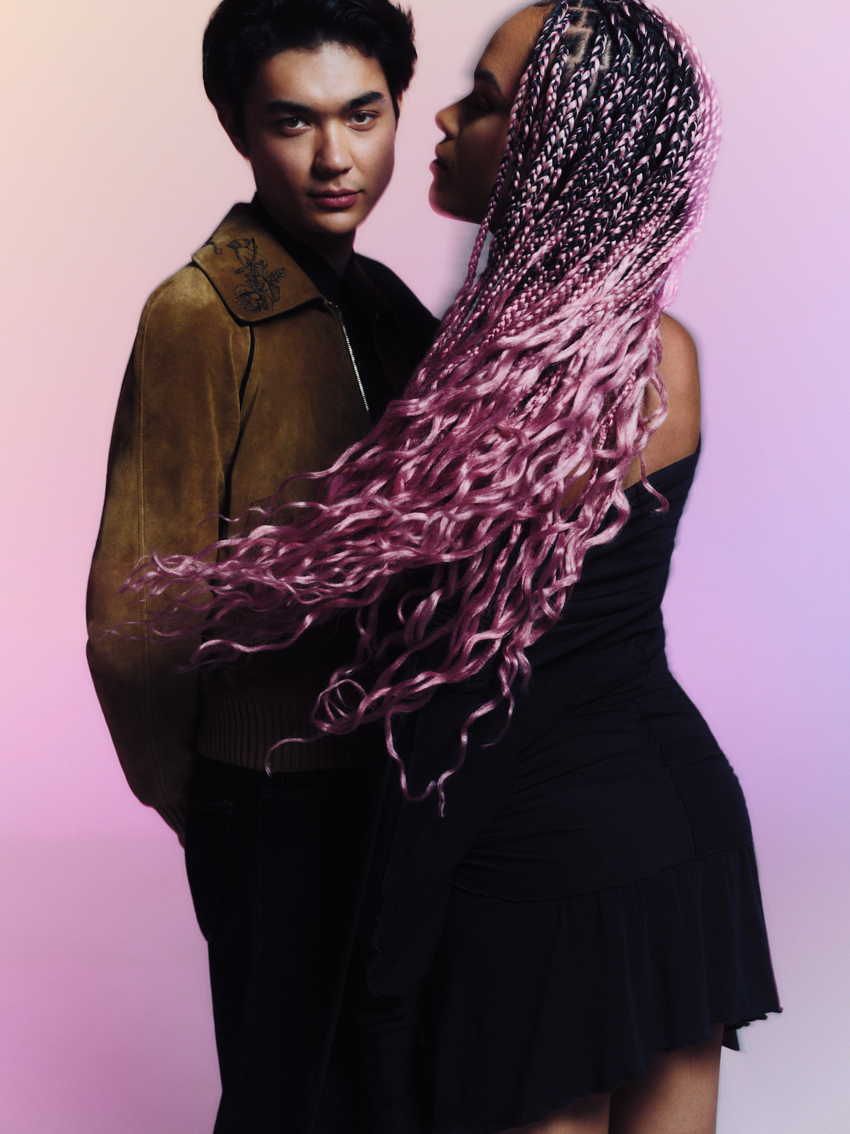 Two people posing together in a studio with soft pastel lighting; one person wears a brown jacket, the other has long pink braids and a black dress, creating a dynamic scene with a stylish, modern atmosphere.