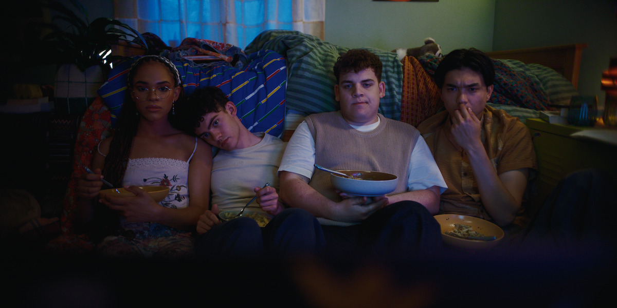 Four friends sitting on the floor in a cozy, dimly lit bedroom, eating food from bowls and watching something together, surrounded by pillows and blankets.