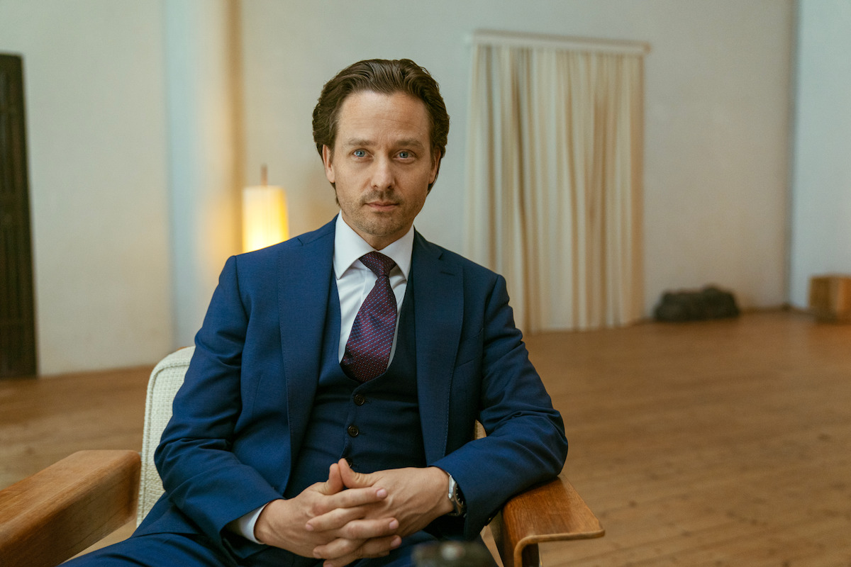 Tom Schilling as Björn in “Murder Mindfully” wears a dark blue three-piece suit and looks smugly into the camera while sitting on a chair with his hands folded.