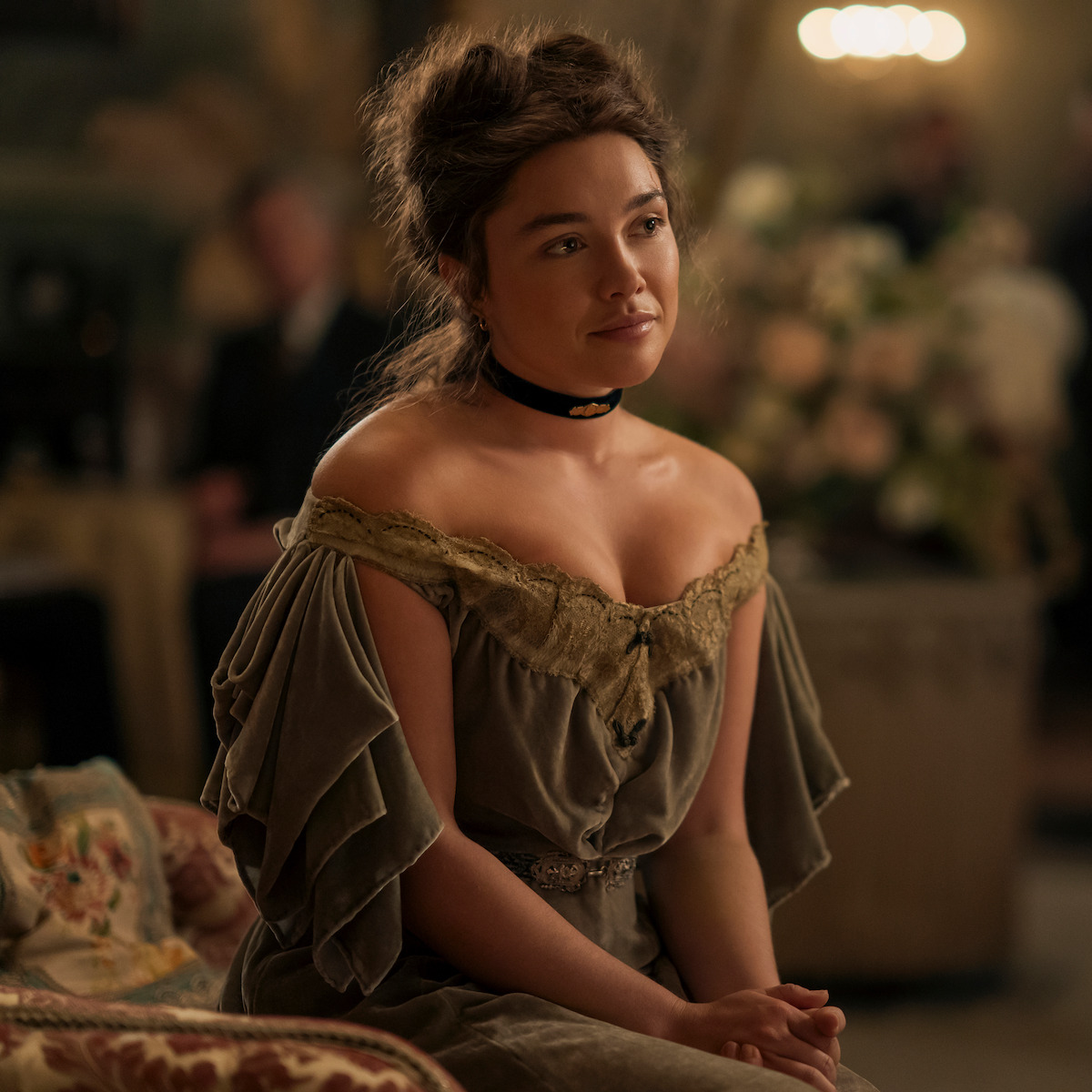 Florence Pugh como Cathy Ames, com um vestido de época de ombros à mostra, sentada em um sofá estampado em um ambiente interno elegante e histórico, com luz suave e um arranjo de flores ao fundo.