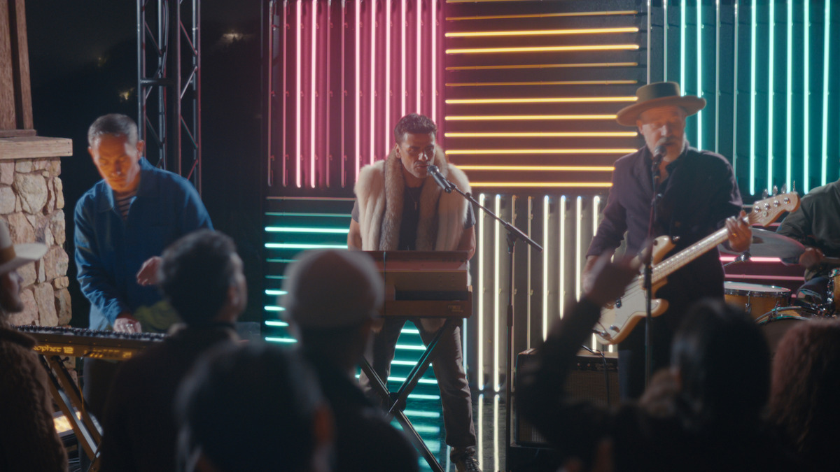 A band performs live on stage with colorful neon lights in the background, including a keyboard player, singer, and guitarist. Audience members are watching and raising their hands in an outdoor nighttime concert setting.