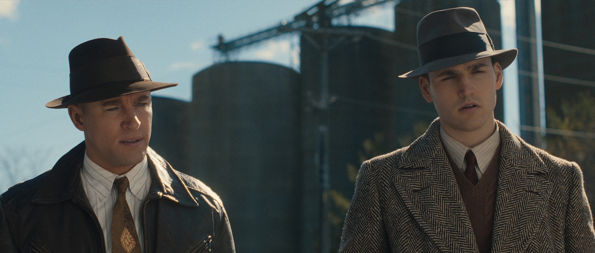 Two men wearing suits and fedora hats stand outdoors in front of large industrial silos, looking serious.
