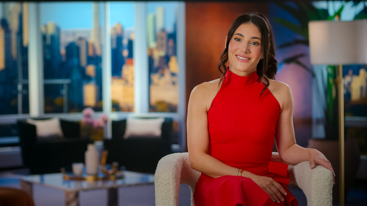 Woman in a red dress sits smiling in a modern apartment with large windows, city skyline view, and stylish decor, creating a bright, welcoming, and elegant atmosphere.