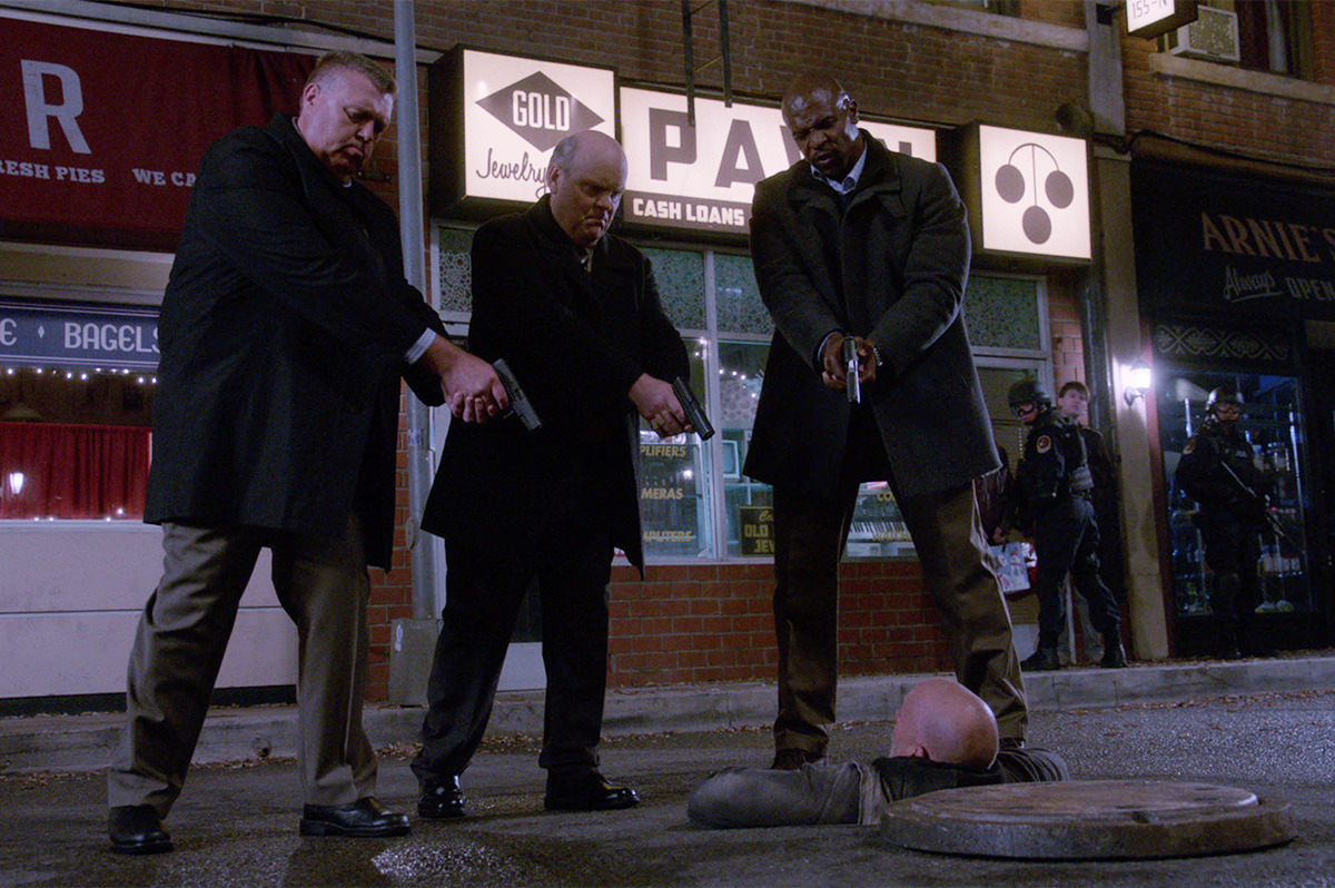 Three men standing with guns, pointing down at another man who is attempting to escape from a manhole.