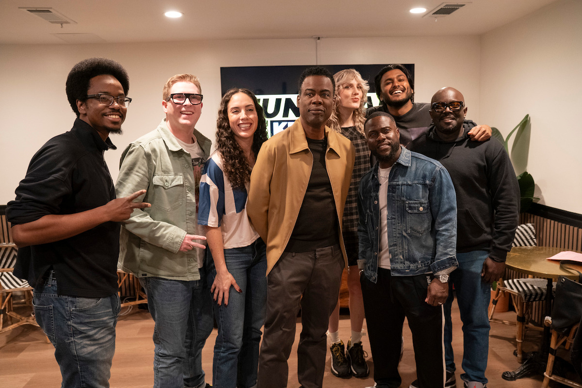 Group of eight people posing indoors in a casual setting with chairs, a table, and a plant in the background; everyone is smiling, wearing casual clothes, and the environment appears friendly and relaxed.
