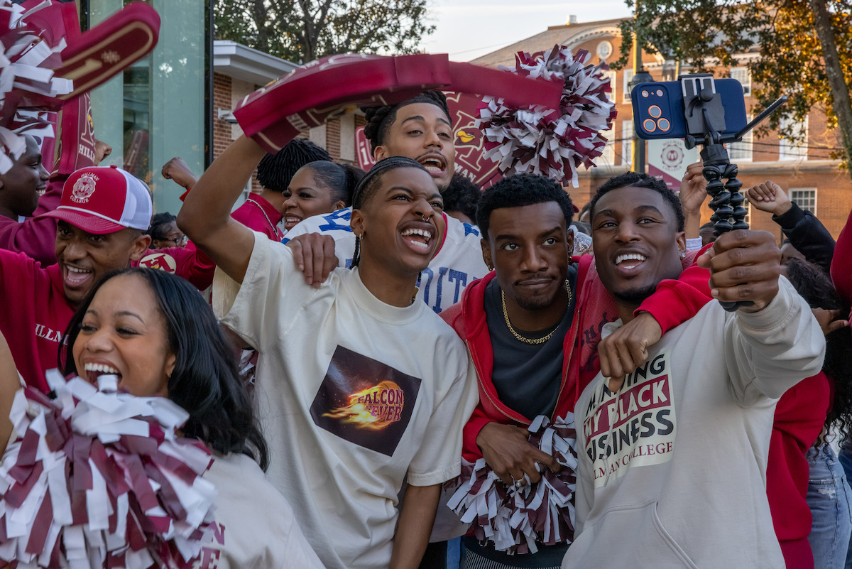 College students at a lively outdoor event, smiling and cheering with pom-poms and team gear, taking selfies and enjoying a festive, energetic atmosphere on campus.