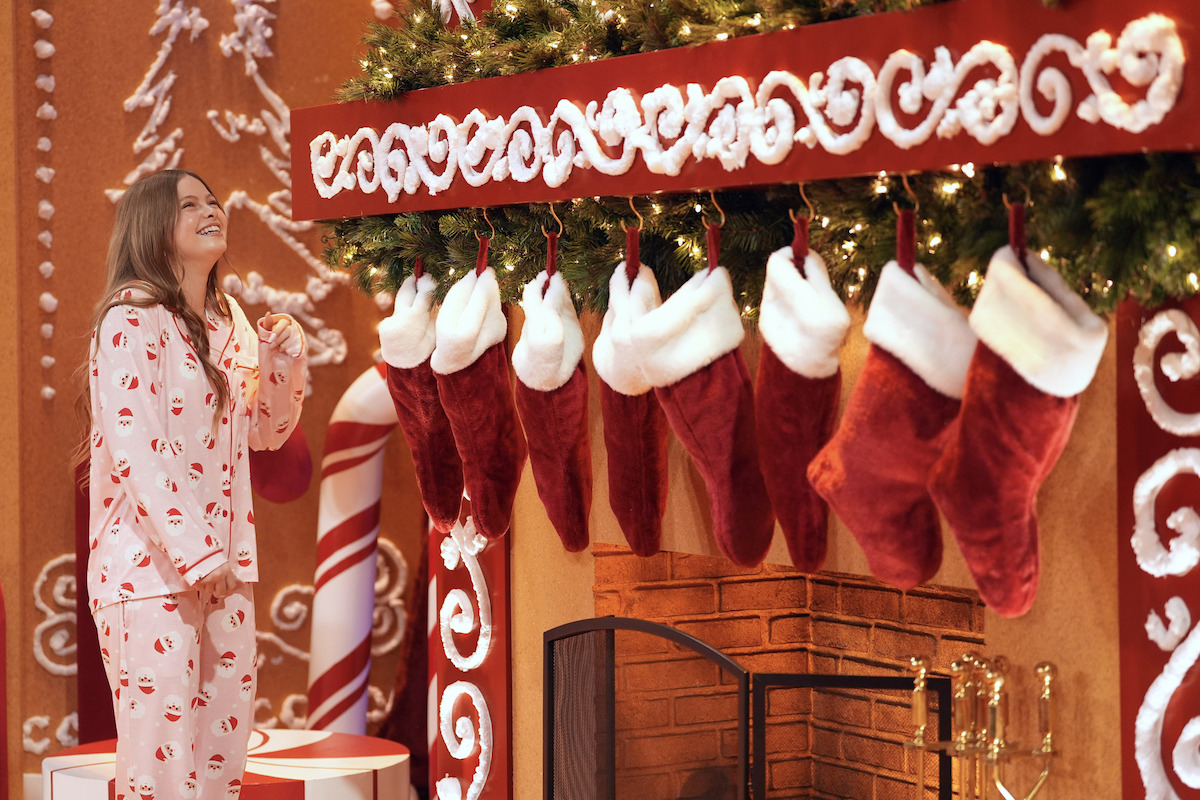 Person in festive pajamas standing by a decorated fireplace with stockings hanging, surrounded by holiday decorations including garlands, candy cane props, and gingerbread-style adornments, creating a warm Christmas scene.