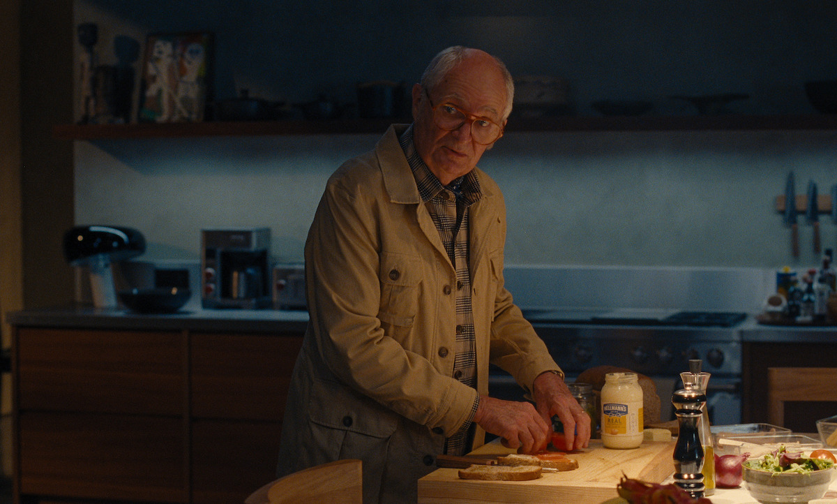 Elderly man in a beige coat preparing food in a modern kitchen, surrounded by ingredients on the counter, with warm lighting creating a cozy, contemplative mood.