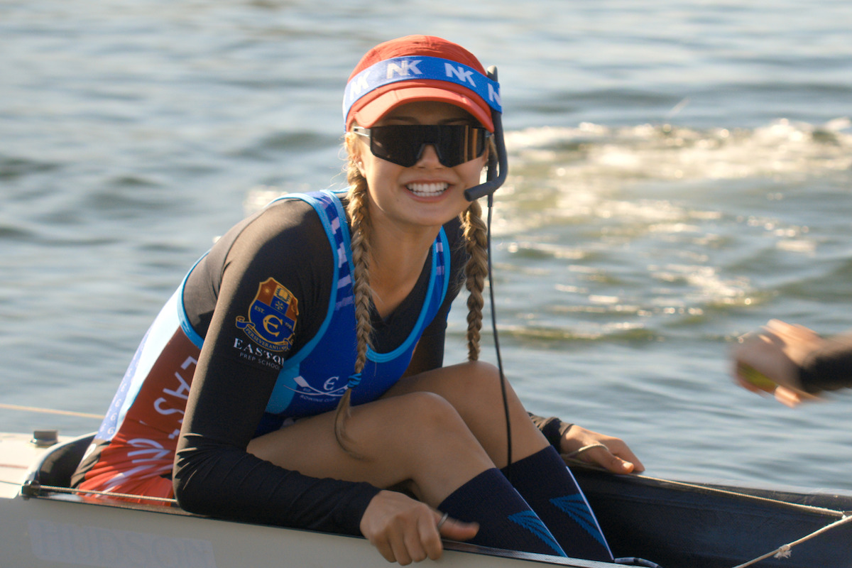 Smiling woman in rowing gear with sunglasses and braids sits in a racing boat on a sunlit body of water, teammate's arm visible, suggesting an outdoor rowing competition or practice.
