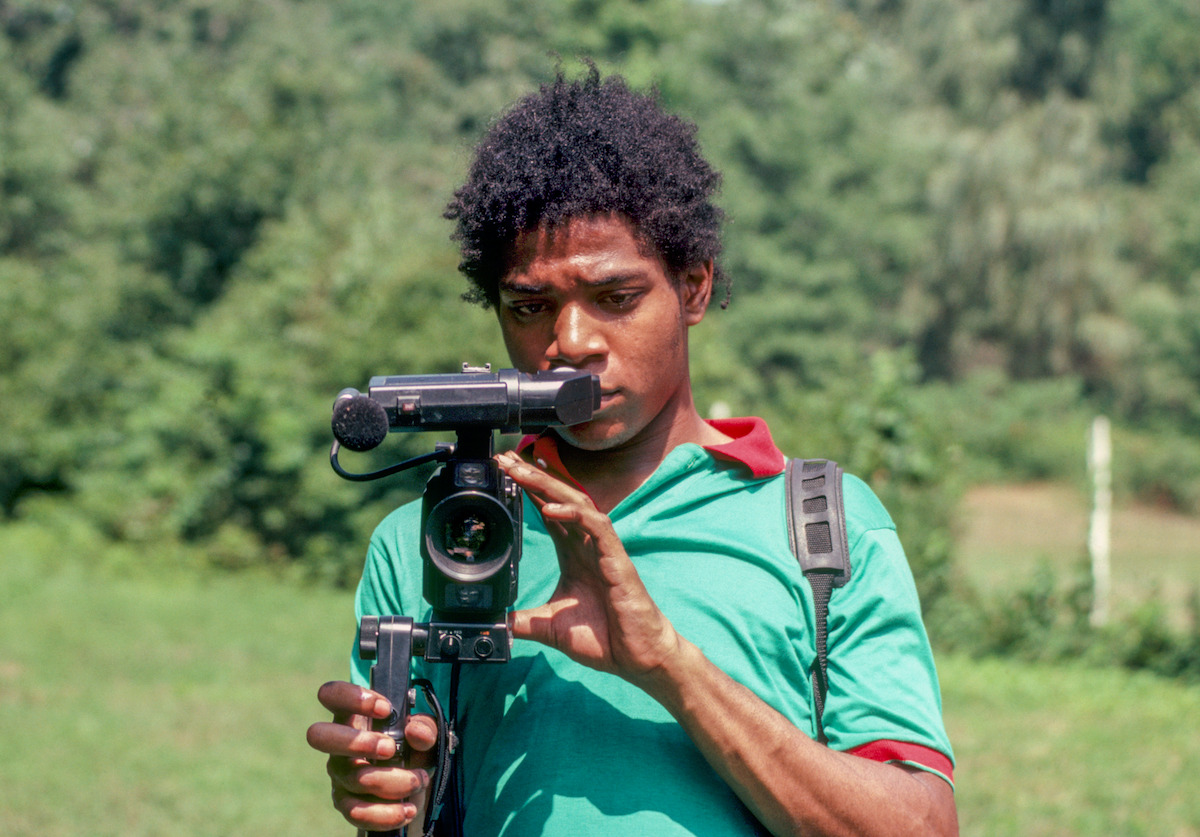 A person outdoors in a green shirt and backpack operating a video camera on a tripod, surrounded by greenery and trees in a natural setting.