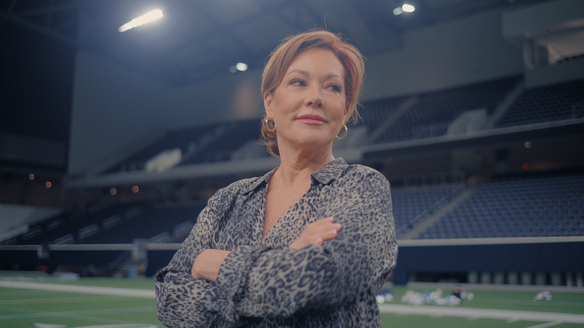 Confident woman with crossed arms stands on an empty indoor sports stadium field, wearing a patterned blouse, with bright stadium lights and empty seating in the background.