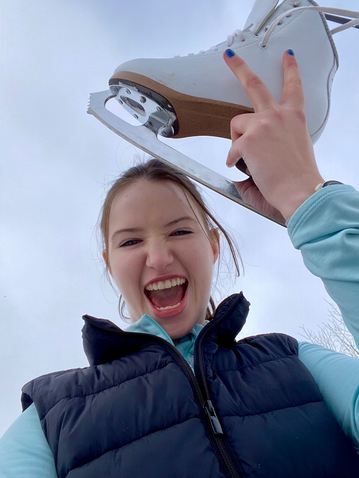 Smiling young woman outdoors in winter attire holding an ice skate above her head, cloudy sky in the background, appearing happy and excited.